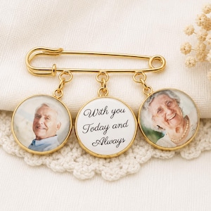 May include: A gold-tone safety pin with three circular pendants. Two pendants display photographs of people, and the central pendant bears the inscription "With you Today and Always". The pin rests on a white, textured surface.