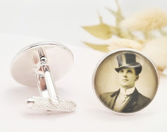 May include: Silver cufflinks with a vintage sepia-toned portrait of a man in a top hat. The cufflinks have a round face and a textured, rectangular bar. The background is a soft, white fabric.