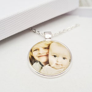 Puede incluir: Collar con colgante plateado con un dije circular con una fotografía en color de dos niños. El colgante cuelga de una fina cadena, presentado en una caja blanca abierta.