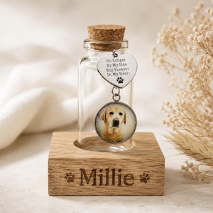 May include: A clear glass memorial bottle with a cork stopper, holding a dog photo charm. The charm has a heart-shaped tag with the text "No Longer By My Side But Forever In My Heart." The wooden base is engraved with the name "Millie."