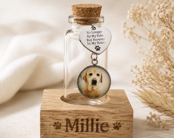 May include: A clear glass memorial bottle with a cork stopper, holding a dog photo charm. The charm has a heart-shaped tag with the text "No Longer By My Side But Forever In My Heart." The wooden base is engraved with the name "Millie."