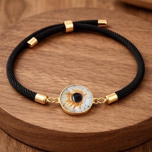 Puede incluir: Una pulsera de cordón negro con un dije circular central que muestra un ojo humano realista. El dije está engastado en un marco de metal dorado, y el cordón está rematado con terminales y anillas de unión de metal dorado a juego.