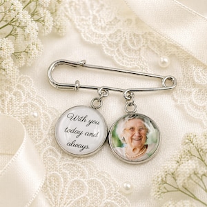 Op de afbeelding: Een zilveren veiligheidsspeld met twee ronde bedels. Eén bedel heeft een foto van een lachende vrouw, de andere de tekst "With you today and always". De speld ligt op een wit kanten oppervlak met parels en bloemen.