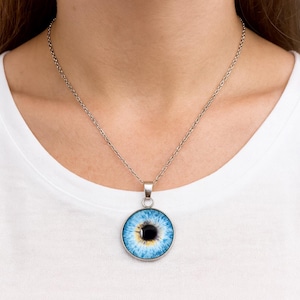 Könnte beinhalten: Eine silberfarbene Halskette mit einem runden Anhänger, der eine realistische Grafik eines blauen menschlichen Auges zeigt. Der Anhänger hängt an einer schlichten silberfarbenen Metallkette und zeigt eine detaillierte Iris in Blau, Weiß und Gelb.