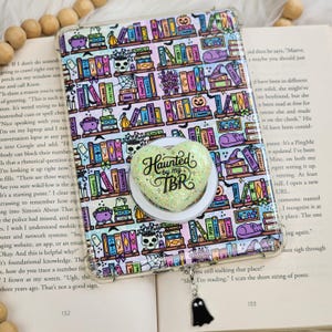 May include: A clear tablet case featuring a colorful pattern of bookshelves filled with books, skulls, pumpkins, and cats. A heart-shaped, glittery phone grip with the text "Haunted by my TBR" is attached, along with a small black ghost charm.