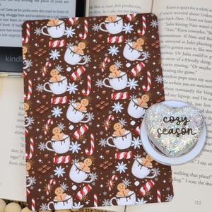 May include: A rectangular brown tablet cover featuring a repeating pattern of gingerbread people in white mugs, red and white candy canes, chocolate squares, and white snowflakes. A heart-shaped white phone grip with glitter and the text cozy season.