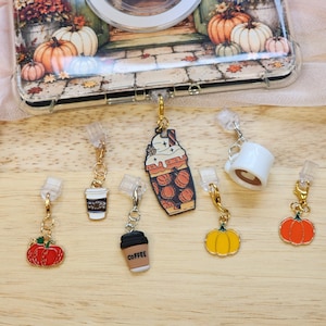 May include: A collection of six metal charms featuring autumn themes, including pumpkins and coffee cups, attached to clear plastic clips. One large charm depicts a pumpkin-patterned drink with whipped cream, while others show individual pumpkins and coffee cups.