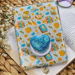 May include: A clear phone case with a whimsical pattern of books, lemons, seashells, and starfish. A blue glitter heart phone grip is attached. A gold charm with a heart pendant hangs from the case.