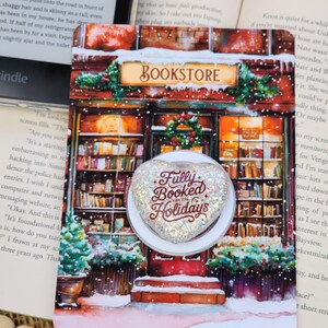 May include: A heart-shaped phone grip with a glittery finish and the text Fully Booked for the Holidays in maroon script. The grip is mounted on a card featuring a festive illustration of a bookstore storefront with snow and greenery.