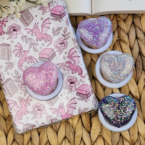 May include: A clear phone case with a pink dragon and book pattern, paired with heart-shaped glitter phone grips in pink, purple, silver, and blue. The grips are mounted on white circular bases, set against a woven background.
