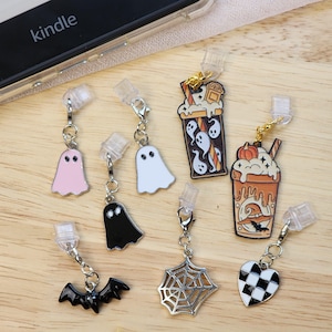 May include: A collection of seven Halloween-themed charms with clear plastic clips. The charms include a pink ghost, a white ghost, a black ghost, a black bat, a silver spider web, a checkered heart, and two beverage cups with ghost and pumpkin designs.
