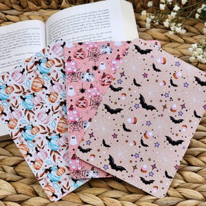 May include: Three rectangular notebooks featuring Halloween patterns. The left notebook has blue and orange pumpkins with leaves. The center notebook displays pink pumpkins, ghosts, and spiderwebs. The right notebook shows black bats, white spiderwebs, and candy corn.