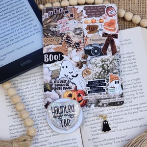 May include: A heart-shaped phone grip with silver glitter and the black text Haunted by my TBR, accompanied by a small black ghost charm, resting on a collage card featuring autumn imagery, pumpkins, ghosts, and text, placed on an open book.