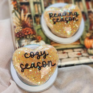 May include: Two heart-shaped phone grips with a translucent orange resin finish filled with iridescent glitter. One grip features the black text "cozy season" and the other features the black text "reading season" in a cursive script font.