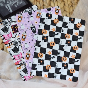 May include: Three rectangular e-reader skins featuring Halloween patterns. One has a black and white checkerboard with orange pumpkins, ghosts, and bats. Another has a light purple background with ghosts, spiderwebs, and coffee cups. The third has books, mugs, and skulls.