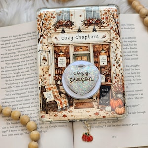 May include: A clear phone case featuring a bookstore illustration labeled cozy chapters, with a heart-shaped glittery pop socket displaying the text cozy season. A small dangling pumpkin charm is attached to the bottom of the case.