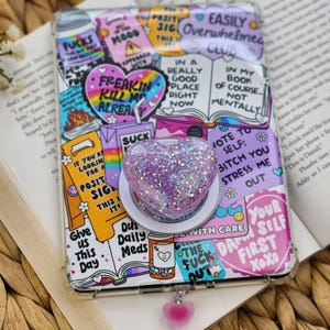 May include: A phone case with a collage of colorful stickers and text, including a heart-shaped pop socket with glitter. The case has a clear border and a pink heart charm. The stickers have phrases like "Freakin Kill Me Alrea" and "Give Us This Day".