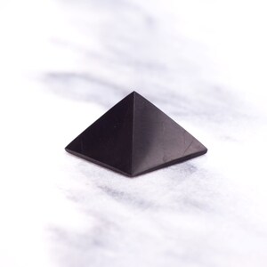 May include: A black obsidian pyramid crystal on a white marble surface.