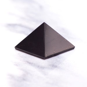 May include: A black obsidian pyramid crystal on a white marble surface.