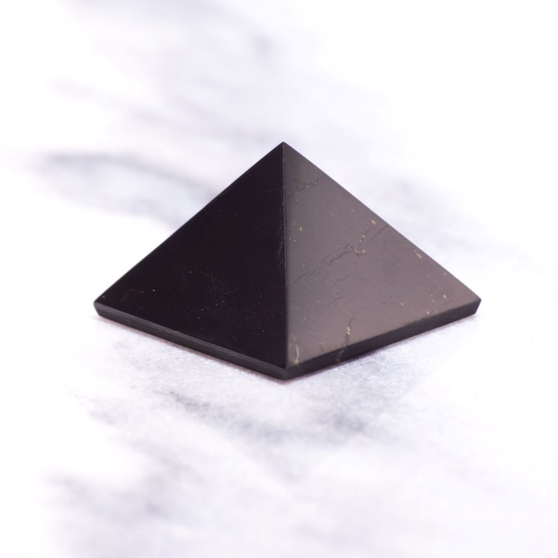 May include: A black obsidian pyramid crystal on a white marble surface.