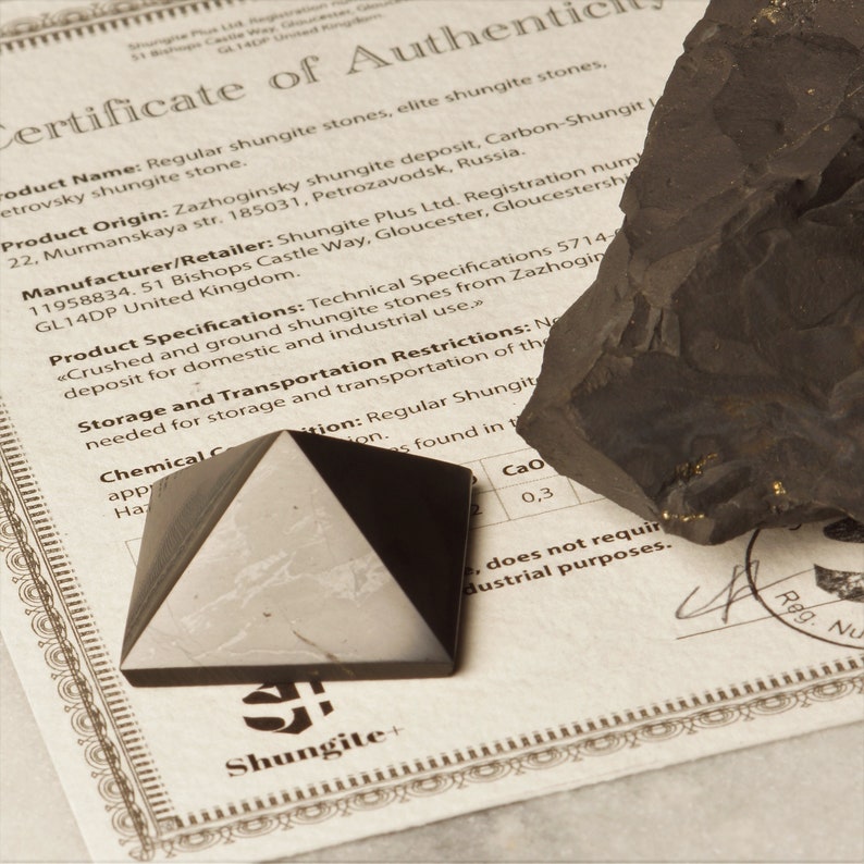 May include: A certificate of authenticity for shungite stones. The certificate is printed on a white paper with black text and a decorative border. The certificate includes the product name, origin, manufacturer, and specifications. The image also shows a black shungite pyramid and a piece of raw shungite.