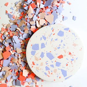 Könnte beinhalten: Ein runder, cremefarbener Untersetzer mit Terrazzo-Design aus blauen, orangefarbenen und beigefarbenen Fragmenten. Die Worte "SOFT CLAY" sind schwarz gedruckt. Passende Fragmente in ähnlichen Farben sind um den Untersetzer verstreut.