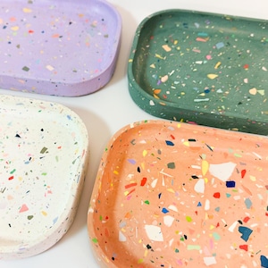 Könnte beinhalten: Vier rechteckige Tabletts mit einem gesprenkelten Terrazzo-Design. Die Tabletts sind in den Farben Lila, Grün, Weiß und Pfirsich.