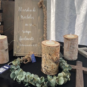 Peut inclure: Une pancarte en bois avec le texte espagnol "El cordon de tres hilos no se rompe facilmente Ecclesiastes 4:12". Trois bougeoirs en écorce de bouleau avec des bougies allumées sont posés sur une surface noire avec une guirlande verte.