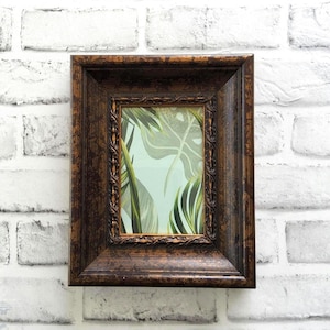 May include: A rectangular brown framed picture with a botanical print of green leaves and a light blue background. The frame has a detailed, ornate design and is mounted on a white brick wall.