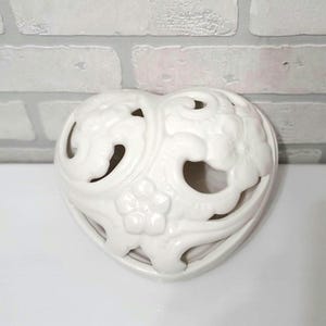 May include: A white ceramic heart-shaped candle holder with floral and swirl cutouts. The decorative piece has a glossy finish and is designed to hold a small candle. The heart is approximately 5 inches wide.