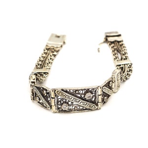 Puede incluir: Un brazalete de plata con un dise&ntilde;o de filigrana y dos peque&ntilde;as piedras transparentes. El brazalete tiene una pieza central rectangular con un patr&oacute;n geom&eacute;trico.