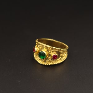May include: A gold ring with a green and red gemstone setting. The ring has an intricate design with a textured surface.