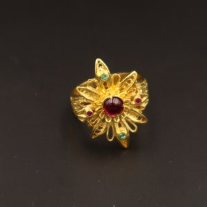 Puede incluir: Un anillo dorado con un diseño floral. El anillo presenta una gema roja grande y redonda en el centro, rodeada de gemas rojas y verdes más pequeñas. El intrincado diseño del anillo es visible sobre un fondo oscuro.