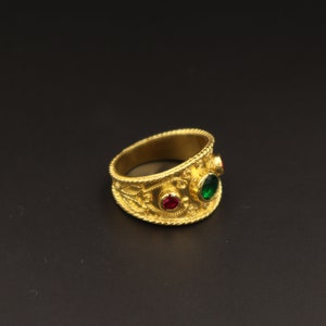 May include: A gold ring with intricate detailing and three gemstones. The ring has a red gemstone in the centre and two green gemstones on either side.