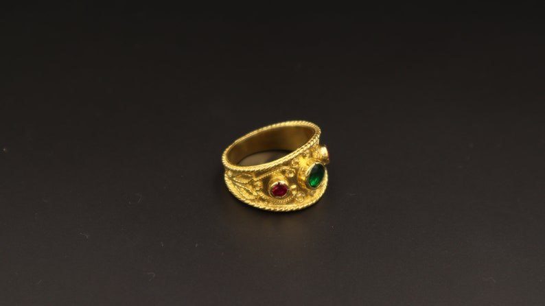 May include: A gold ring with intricate detailing and three gemstones. The ring has a red gemstone in the centre and two green gemstones on either side.