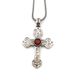 Peut inclure: Un collier en chaîne argent avec un pendentif en forme de croix argent et or. La croix est ornée d'une pierre précieuse rouge au centre et de motifs floraux complexes.