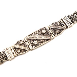 Puede incluir: Un brazalete de plata con una cadena decorativa y una pieza central rectangular con filigrana intrincada y peque&ntilde;as piedras transparentes.