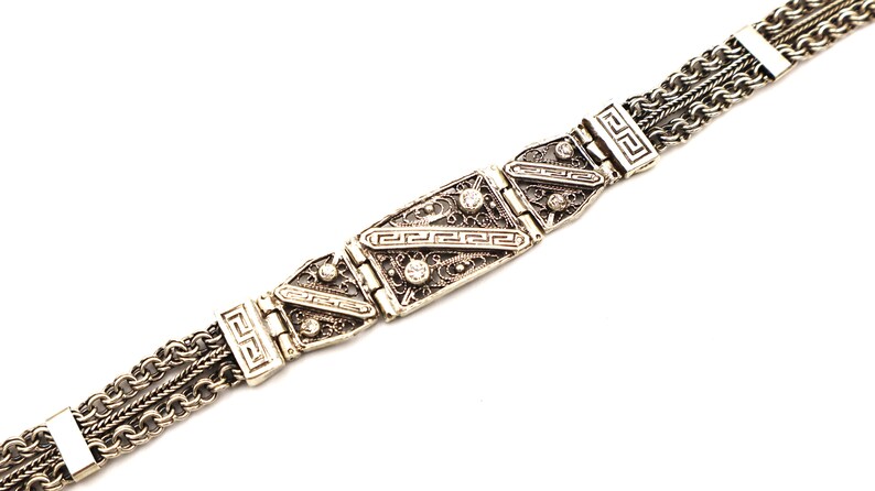 Puede incluir: Un brazalete de plata con una cadena decorativa y una pieza central rectangular con filigrana intrincada y peque&ntilde;as piedras transparentes.