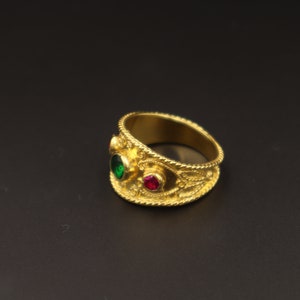 May include: A gold ring with intricate detailing and two gemstones. The larger gemstone is green, and the smaller gemstone is red.