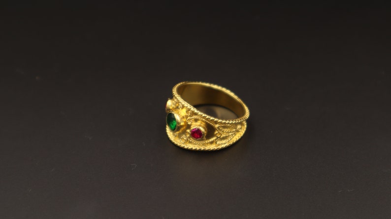 May include: A gold ring with intricate detailing and two gemstones. The larger gemstone is green, and the smaller gemstone is red.