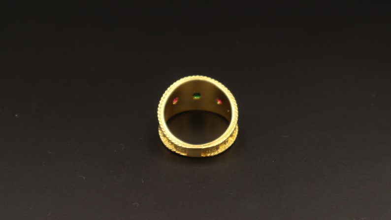 May include: Gold ring with three small gemstones, one green and two red, set into the band.