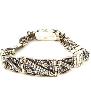 Puede incluir: Un brazalete de plata con una cadena delicada y dise&ntilde;os geom&eacute;tricos intrincados. El brazalete presenta peque&ntilde;as piedras transparentes engastadas en el metal.