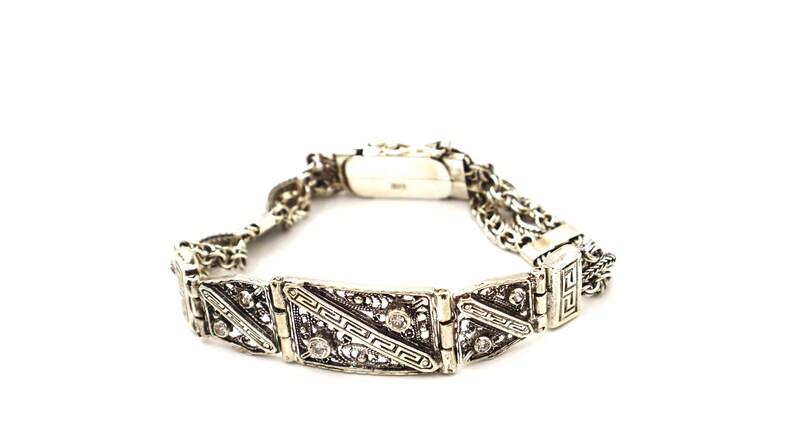 Puede incluir: Un brazalete de plata con una cadena delicada y dise&ntilde;os geom&eacute;tricos intrincados. El brazalete presenta peque&ntilde;as piedras transparentes engastadas en el metal.