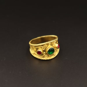 May include: A gold ring with intricate detailing and three gemstones. The ring has a green gemstone in the centre and two red gemstones on either side.
