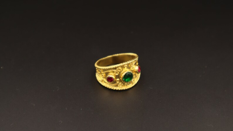 May include: A gold ring with intricate detailing and three gemstones. The ring has a green gemstone in the centre and two red gemstones on either side.