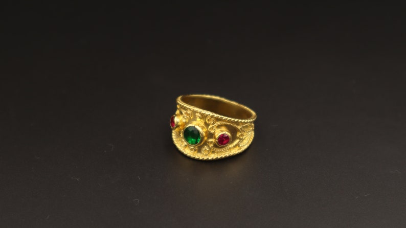 May include: A gold ring with a green and red gemstone setting. The ring has an intricate design with a textured surface.