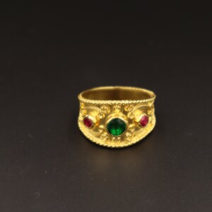 May include: A gold ring with a wide band featuring an intricate design and three gemstones. The centre stone is green, and the two smaller stones on either side are red.