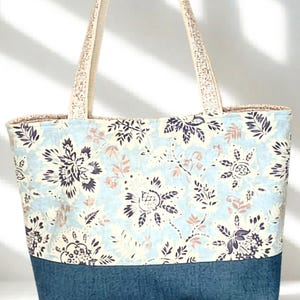 May include: A tote bag featuring a light blue background with a dark blue and tan floral pattern on the upper portion and a solid dark blue textured base. The bag includes two matching light-colored fabric shoulder straps at the top.