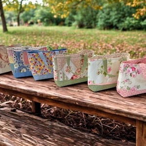 May include: A row of six quilted fabric tote bags featuring various floral, gingham, and patchwork patterns in shades of pink, blue, green, and cream. Each bag has two matching fabric handles and a structured rectangular base.