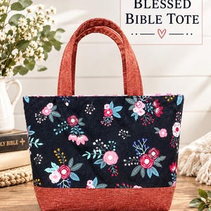 May include: A black tote bag with a floral pattern featuring pink, red, and blue flowers and green leaves. The bag has a textured, rust-colored base and matching handles. Text on the image reads: The BLESSED BIBLE TOTE and Aubrey Creations.